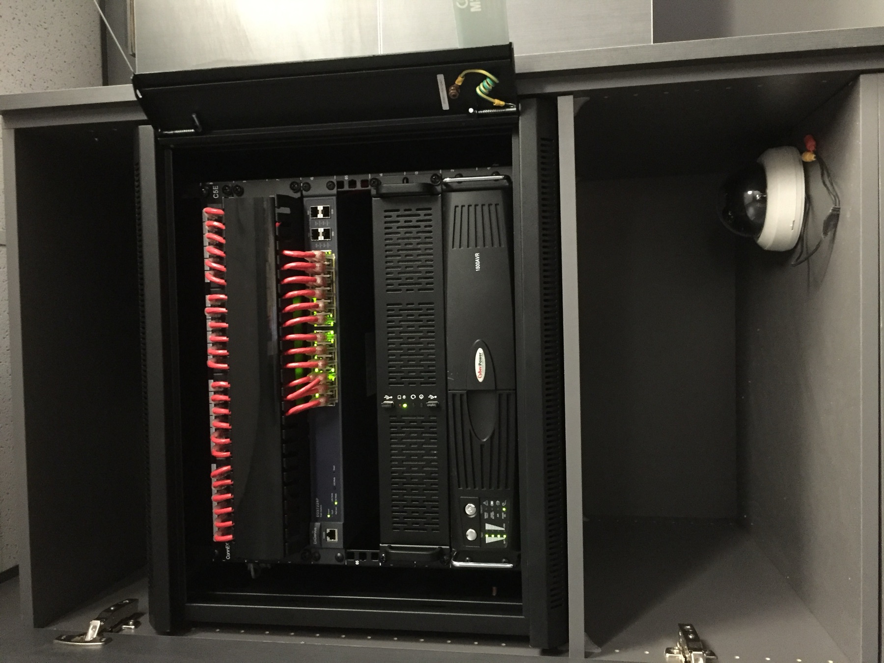 Grey cabinet rack with patching, UPS, and a staged dome camera.