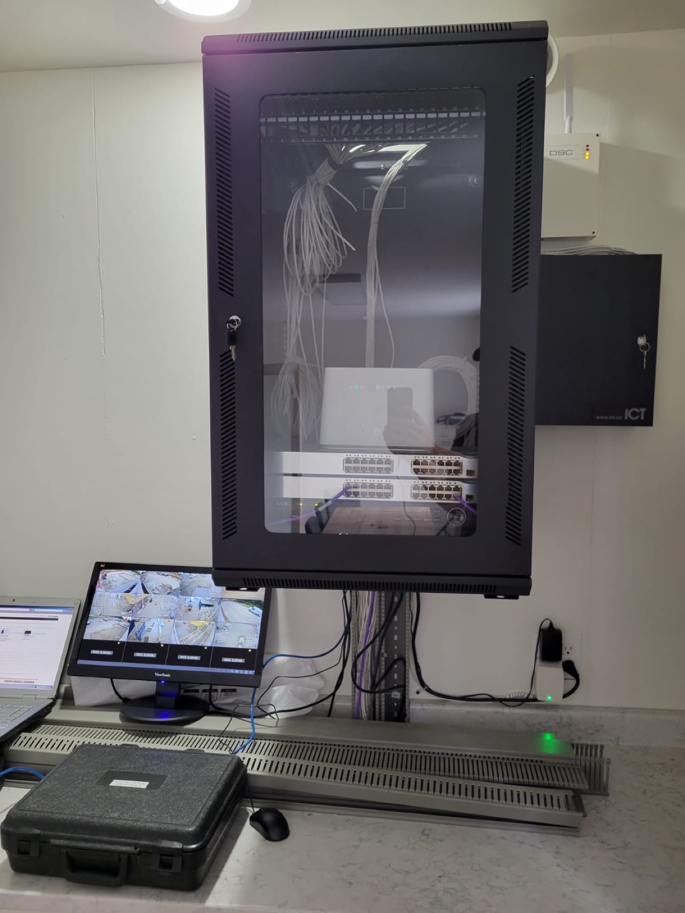 Glass-front wall rack, ICT enclosure, and a desk monitor.